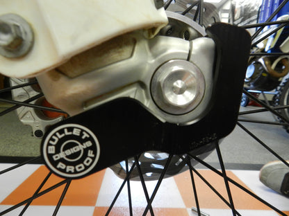 A black Bullet Proof Designs fork lug guard designed to fit on a motorcycle's front suspension system.